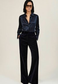 Vrouw in een donkerblauwe satijnen button-up shirt met brede kraag, bijpassende hooggesneden fluwelen wijde broek en donkere zonnebril, met handen in de zakken.