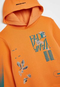 Sudadera con capucha naranja que presenta un gráfico frontal con la palabra "FADE" en azul, un estampado de hojas y detalles de texto. Material de algodón suave con un acabado texturizado.