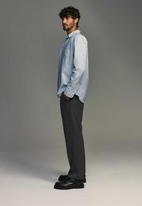 Camisa de botonadura a rayas en azul claro y blanco, combinada con pantalones oscuros de corte entallado en gris y zapatos de cuero negros, de pie contra un fondo neutro.