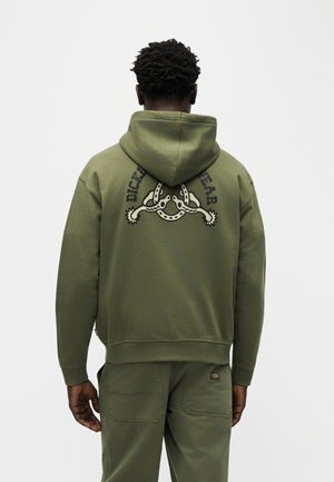 Man draagt een olijfgroene hoodie met een zwarte en beige hoefijzer- en waardesporenafbeelding en de tekst "Dickies Wear" op de achterkant, gecombineerd met bijpassende broek.