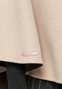 Kangas villa beige valuu pehmeästi. Helman vaaleanpunaisessa merkillä lukee "Käsintehty". Alla mustassa kankaassa on pinstripe-kuvio.
