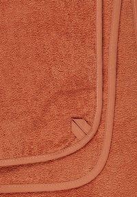 Terracotta towel with a textured, looped surface and a contrasting smooth fabric trim. The edges are finished with neat stitching.
