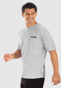 Grå t-shirt med korte ærmer lavet af blødt stof, med rund halsudskæring og sort "SMILODOX" logo på venstre bryst. Modellen har sorte bukser på.