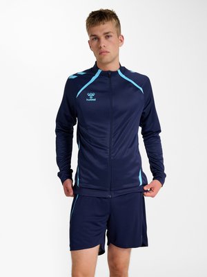 Ung mand iført en marineblå og lyseblå sportjakke og shorts, stående mod en ensartet hvid baggrund.