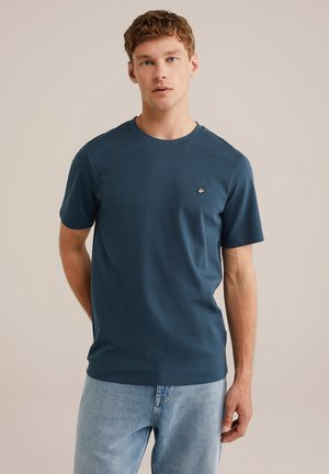 T-shirt a maniche corte di un verde scuro in tessuto morbido, con collo rotondo e un piccolo logo ricamato sul petto. Indossato con jeans chiari.