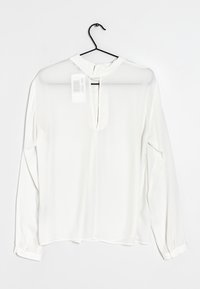 Blusa bianca a maniche lunghe, in tessuto trasparente, design senza colletto, apertura a chiave frontale, maniche con polsino e dettaglio a bottone, appesa a un gancio nero.