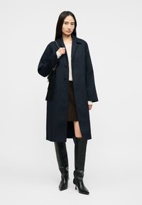 Γυναίκα που φοράει σκούρο μπλε παλτό με κουμπιά, μαύρες μπότες μέχρι το γόνατο, καφέ φούστα και κρατάει μαύρη τσάντα ώμου με κρόσσια.