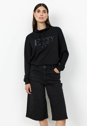 Frau trägt ein schwarzes langärmeliges Oberteil mit dem Schriftzug „Every moment matters“ und schwarze weite kurze Hosen, posiert mit einer Hand in der Tasche.