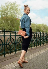 Denim zkrácená bunda, černá pouzdrová sukně, hnědé sandály na podpatku. Malá oranžová kabelka se zlatými detaily, stojící u zábradlí.
