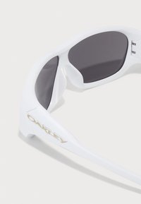 Óculos de sol desportivos brancos Oakley com lentes curvadas escuras e logotipo no braço da armação, apresentados sobre um fundo branco.