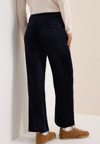 Marineblaue Cordhose mit weitem Schnitt, ausgestattet mit Gesäßtaschen und einer strukturierten Oberfläche, kombiniert mit hellen Schuhen und Socken.