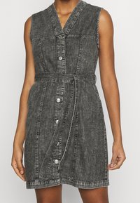 Sleeveless denim dress in faded black with a button-down front, side pockets, and a tie belt. Features contrasting white stitching and a structured shape.