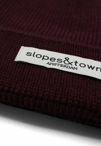 Berretto di maglia bordeaux con un motivo testurizzato. Presenta un'etichetta bianca con "slopes&town AMSTERDAM" in lettere nere.