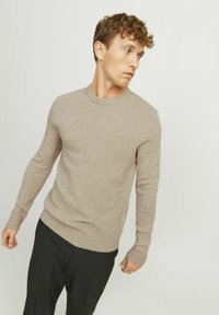 Pull en tricot beige avec un col rond, des poignets et un ourlet côtelés, présentant une texture lisse et une coupe ajustée, associé à un pantalon noir.