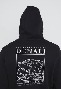 Felpa nera con cappuccio caratterizzata da una grafica bianca di montagne sul retro, con il testo "LA FACCIA NORD DEL DENALI" e dettagli sulla posizione sottostanti.