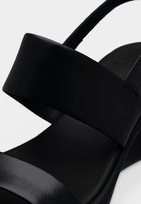 Μαύρο πέδιλο πλατφόρμα με φαρδιές δερμάτινες λωρίδες και τακούνι με ιμάντα γύρω από τη φτέρνα σε απλό λευκό φόντο.