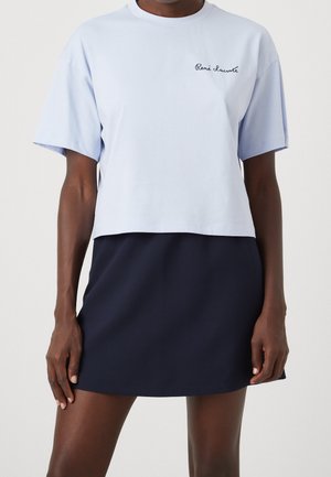 Kvinde iført lyseblå kort T-shirt med teksten "René Lacoste" og en mørkeblå nederdel, stående mod en ensfarvet baggrund.