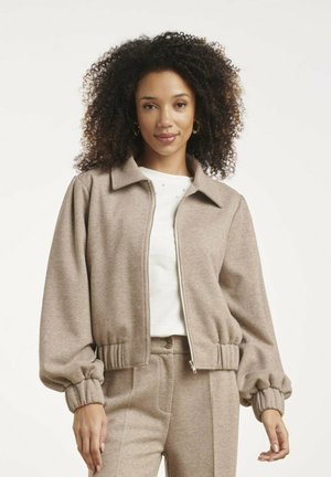 Vrouw met krullend haar die een beige jas met rits en bijpassende broek draagt over een wit shirt, staand tegen een lichte achtergrond.