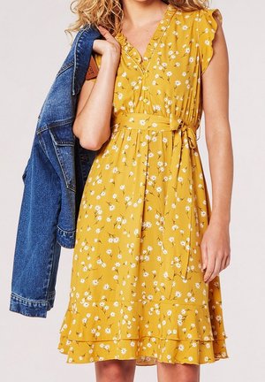 Frau trägt ein gelbes Blumenkleid mit Rüschen und einer Taillenschleife, hält eine blaue Jeansjacke über der Schulter.