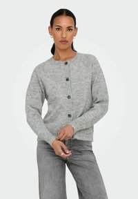 Grå stickad cardigan med rund halsringning, knappframsida och ribbade ärmslut. Modellen bär den med grå denim, med händerna placerade vid midjan.