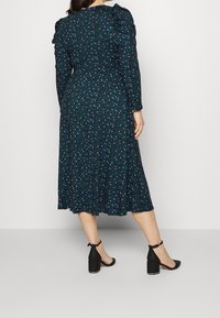 Robe fleurie vert foncé avec manches longues, épaules froncées et ourlet à hauteur des genoux. Accompagnée de talons noirs à bride à la cheville.