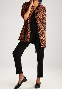 Nainen, joka on pukeutunut leopardikuosiseksi takiksi mustan asun päällä, mustiin lyhytlahkeisiin housuihin ja kiiltäviin mustiin loafer-kengissä, poseeraa yksi käsi kasvojen lähellä.