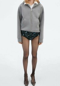 Sweat à capuche gris zippé, short noir à sequins, collants noirs transparents et talons ornés. Présente une coupe décontractée et une texture douce.