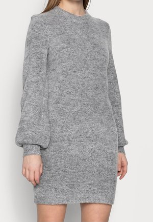 Femme portant une robe pull en maille grise à manches longues avec des manches bouffantes et des poignets côtelés, debout devant un fond clair uni.