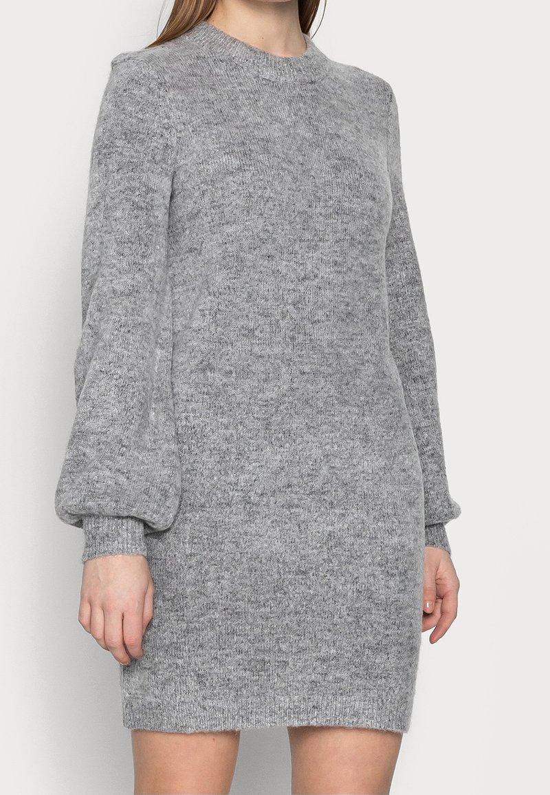 Femme portant une robe pull en maille grise à manches longues avec des manches bouffantes et des poignets côtelés, debout devant un fond clair uni.