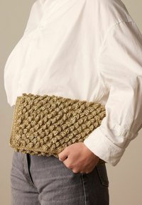 Geweven clutch gemaakt van gestructureerd gouden materiaal, met een driedimensionaal gevlochten ontwerp, gehouden tegen een wit shirt en grijze jeans.