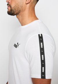 Camiseta blanca de manga corta hecha de algodón, con un logo negro de Puma en el pecho y una franja de acento negra con el nombre de la marca en la manga.