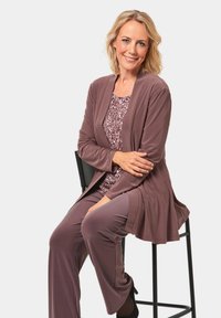 Lange mauve cardigan over een gemusterde lichtpaarse top, gecombineerd met bijpassende broek en zwarte hoge hakken, zittend op een kruk.