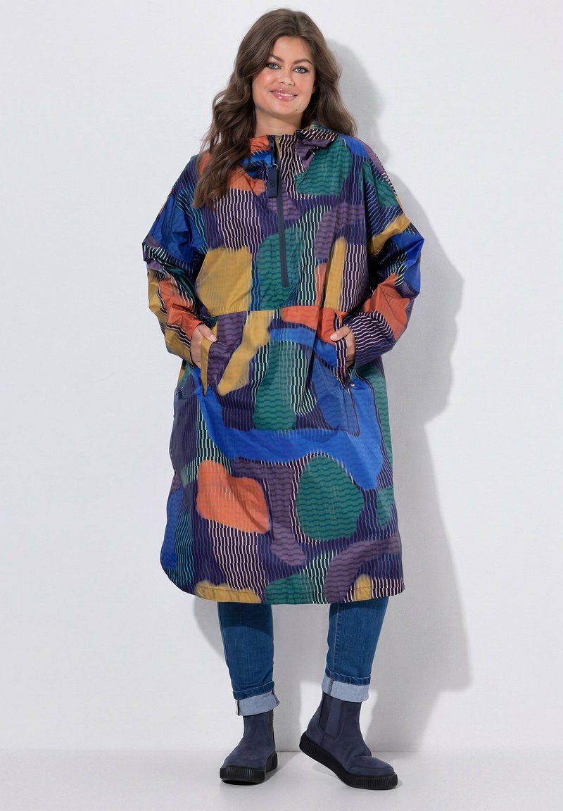 Woman wearing a colorful abstract patterned oversized jacket, blue cuffed jeans, and dark blue ankle boots standing with hands in pockets.