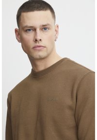 Sudadera de algodón marrón con cuello redondo, puños acanalados y un pequeño logo en el pecho. La tela tiene una textura suave y lisa.