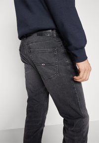 Persoană îmbrăcată cu blugi negri decolorati, cu logo pe buzunarul din spate și un tricou cu mâneci lungi bleu închis, stând în fața unui fundal deschis.