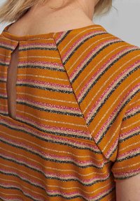 Pruhovaný oranžový top s růžovými, černými a metalickými nitěmi; má otvor ve tvaru klíčové dírky na zádech; žebrovaná textura.