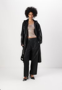 Palton lung din piele sintetică neagră, top din satin metalic bej, pantaloni negri largi și tocuri negre. Accesoriile includ o geantă mică neagră.