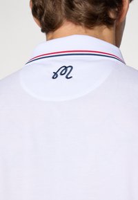 White polo shirt featuring a navy embroidered logo on the back collar, accented with red and navy stripes along the collar edges.
