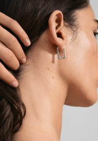 Silver rectangular hoop earring hangs from the ear. Smooth metal surface with a minimalistic design. Model's hand gently touches their hair.