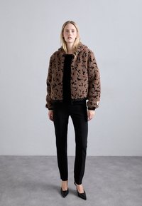 Brown faux fur jacket with black abstract pattern, zip closure, hood, and elastic hem; paired with black fitted pants and pointed black shoes.