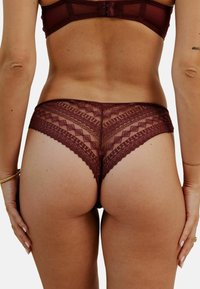 String en dentelle bordeaux avec un motif géométrique et des bords festonnés, présentant une texture douce et un design taille basse. Vue arrière.