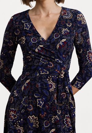 Femme portant une robe portefeuille bleu marine avec des motifs floraux rouges, blancs et bleus, manches trois-quarts, nouée à la taille.