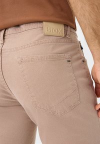 Brax tan denim jeans met een gestructureerde afwerking, voorzien van een leren merkpatch, achterzakken met stikdetails en een slim fit ontwerp.
