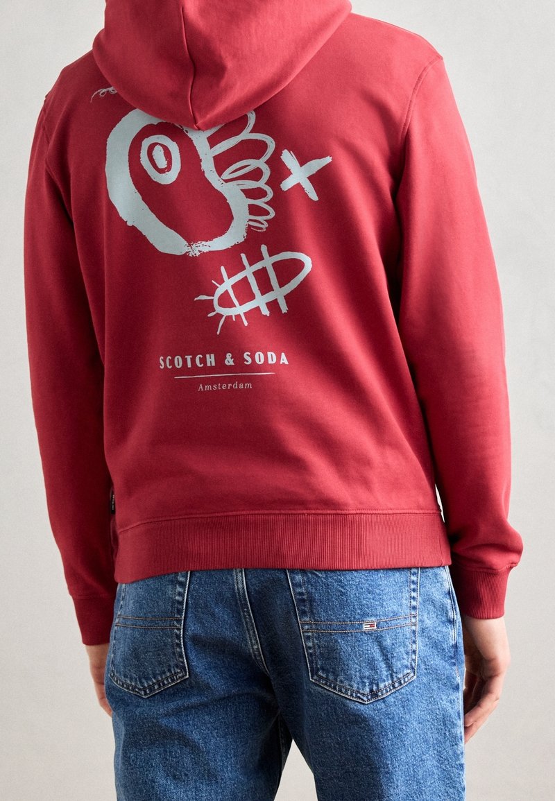 Felpa rossa con un design grafico blu che presenta un pesce stilizzato e testo. Il materiale è morbido, con polsini e orlo a coste. Indossata con jeans blu.