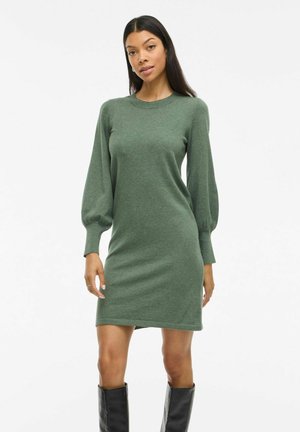 Robe en tricot verte avec un col rond, des manches bouffantes longues et une silhouette ajustée, associée à des bottes noires jusqu'aux genoux.