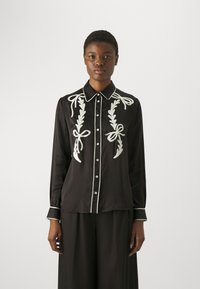 Blusa nera con ricami bianchi, caratterizzata da disegni di fiocchi e foglie. Ha un colletto, apertura con bottoni e Piping bianco a contrasto lungo i bordi.