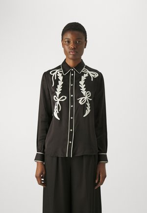 Blusa nera con ricami bianchi, caratterizzata da disegni di fiocchi e foglie. Ha un colletto, apertura con bottoni e Piping bianco a contrasto lungo i bordi.