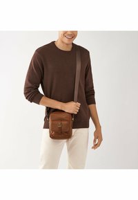 Sac en cuir marron porté en bandoulière avec un rabat avant, détail de boucle et bandoulière réglable, tenu par une personne vêtue d'un pull noir.