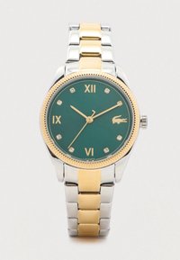 Montre bicolore or et argent avec un cadran vert. Présente des index horaires dorés, des accents en diamant et une lunette texturée en or. Logo de la marque affiché.