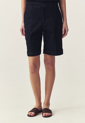 Person trägt dunkelblaue Denim-Bermudashorts mit umgeschlagenen Säumen und schwarze Slide-Sandalen, steht vor einem einfarbigen Hintergrund.